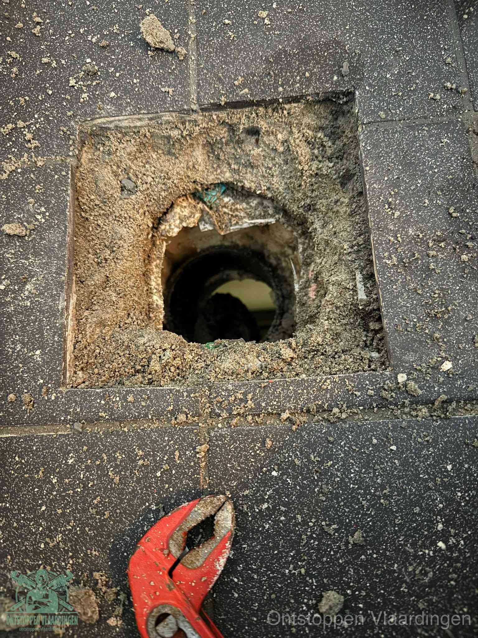 Een geopend rioolputje in een betontegelvloer met losgemaakt rooster en een zichtbare ronde afvoerbuis, het achterhalen waar de afvoerverstopping zit in Vlaardingen