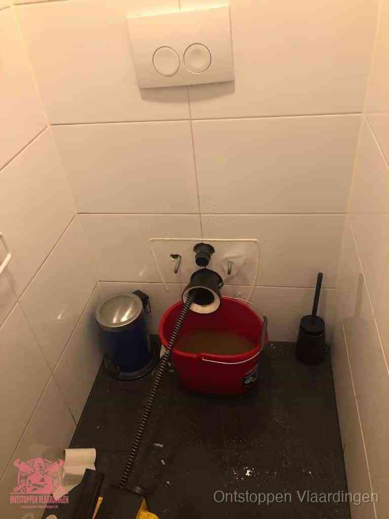 Een ontstopte wand-wc met verwijderde toiletpot waar een ontstoppingsveer in de wanduitgang verdwijnt, het achterhalen waar de verstopping zit in Vlaardingen