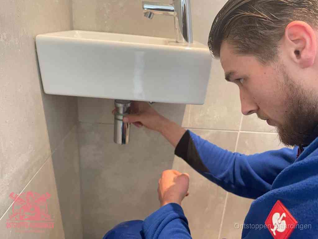 Een monteur draait met de hand de zwanenhalssifon onder een fonteinwasbak in de toiletruimte los om de bron van rioollucht in Vlaardingen te vinden