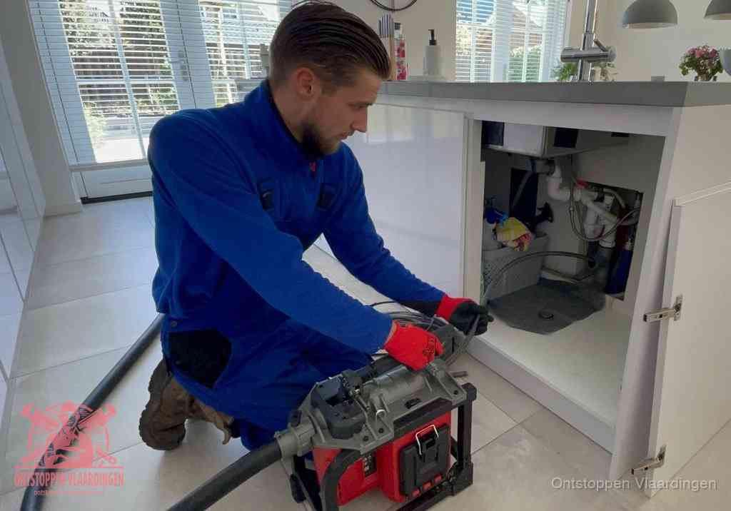 Een monteur voert een professionele elektrische ontstoppingsmachine in de afvoer onder de spoelbak tijdens het gootsteen ontstoppen in Vlaardingen